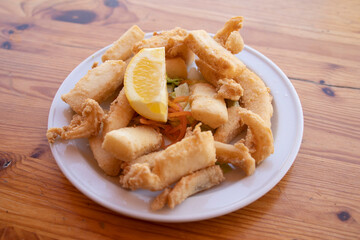 Fried cuttlefish strips, called Chocos Fritos in Spanish. It is served on a white plate on a wooden table. Spanish food concept.