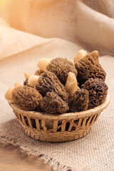A wicker basket with morel mushrooms stands on a burlap cloth lying on a brown wooden background, vertical photo