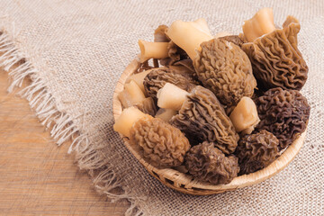 A wicker basket with morel mushrooms stands on a burlap cloth lying on a brown wooden background, top view