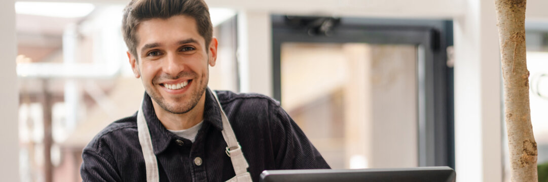 Smiling young man in apron standing at the cash register