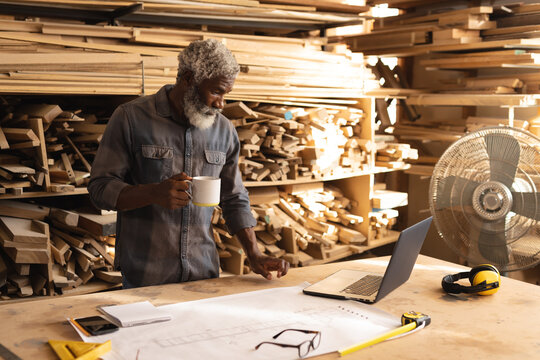 African american mature carpenter working on laptop while having coffee in woodworking workshop