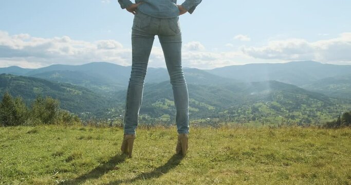 Legs of a woman in jeans and high heels, with stunning views of the mountains and the valley. Blue sky, white clouds, medium shot, daytime, spring, summer