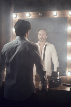 Man in white unbuttoned shirt with balck tie in front of mirrow with lights looking at own reflection