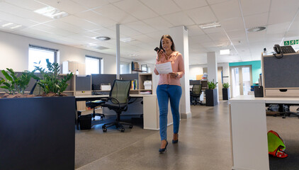 Full length of biracial businesswoman walking with document while talking on smartphone at workplace - Powered by Adobe