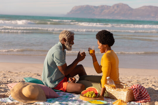 African american couple toasting juice while sitting with food on blanket against sea at sunset