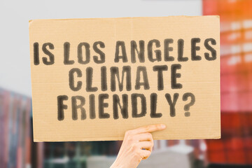 The question " Is Los Angeles climate-friendly? " is on a banner in men's hands with blurred background. Support. Team. Activist. Urban. Sunset. Carbon. Ecology. Energy. New. Clean. Warming. Waste