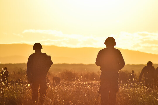 Dos soldados caminan por una pradera con monta&ntilde;as durante el atardecer. Camaradas militares con ropa de guerra caminan a contraluz y dejan ver su silueta. Usan casco, portan equipo y arma