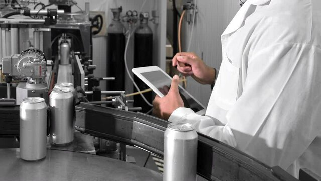Male worker types on a tablet near conveyor with cans at beverage factory production line, close up. High quality 4k footage
