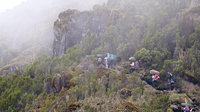 The general plan of porters and tourists are walking along the slope of a rocky mountain during a hike. A chain of porters with equipment walking along a narrow path while climbing Kilimanjaro.