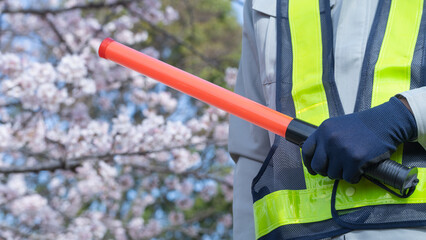 桜の背景で作業服で誘導棒を持つ人｜建設業・警備業