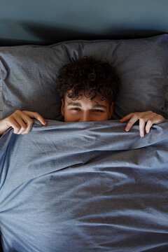 Teen boy hiding under blanket while lying on bed
