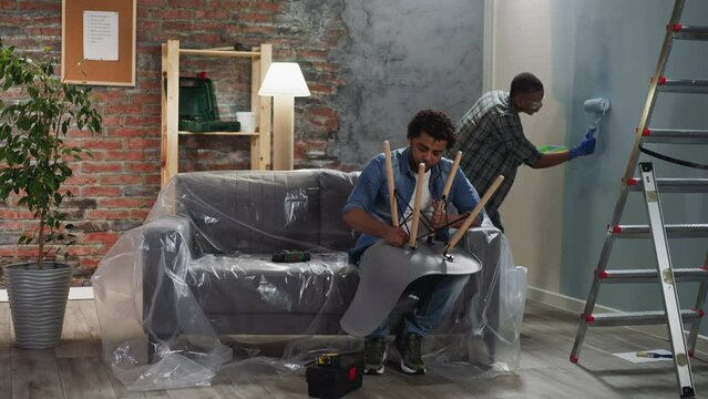 African-American guy paints wall while brother assembles chair in living room. Black friend do domestic repair together. Reconstruction of apartment