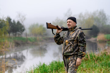 A handsome man with a gun on his shoulder looks at the lake in the fog. Medium plan