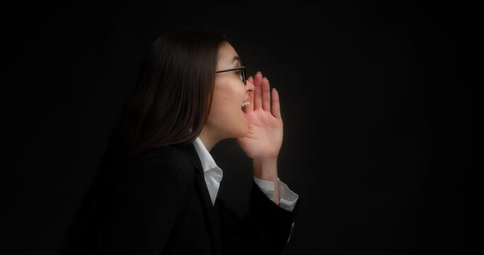 Business woman in glasses and office clothes stands in profile against an isolated black background, looking for you, presses her hand to her mouth, screams loudly.Hey, where are you