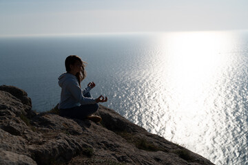 Fototapeta premium Woman tourist enjoying the sunset over the sea mountain landscape. Sits outdoors on a rock above the sea. She is wearing jeans and a blue hoodie. Healthy lifestyle, harmony and meditation