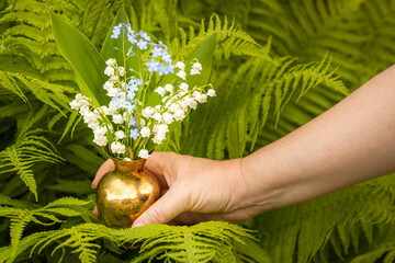 Lilies of the valley in May and forget-me-nots in a charming little jug held in a woman's hand against the background of a garden and green ferns, Spring flowers