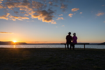 Fototapeta na wymiar Couple sitting on a bench by Lake Rotorua and watching sunset, Rotorua, New Zealand.