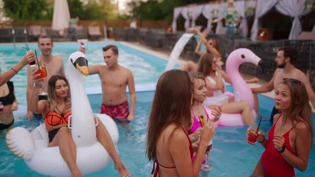 People have swimming pool party with cocktails at a luxury resort. Friends in swimwear enjoying drinks, hanging out and clubbing with inflatable floats. Hot girls relax in the water. Slow motion.