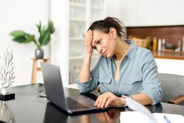 Tired and worn out from online work woman sitting in front laptop holding head, feels strong headache. Female freelance is massaging head with suffering face expression