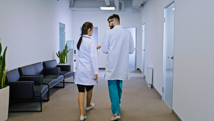 In a modern hospital corridor walking in front of the camera two doctors female and man they have a discussing about some problems of the patient background other hospital stuff take a sit on the