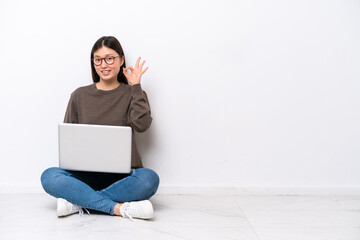 Young woman with a laptop sitting on the floor showing ok sign with fingers