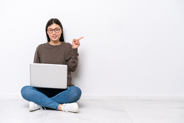 Young woman with a laptop sitting on the floor pointing finger to the side