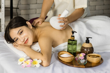 Asian young beautiful spa customer laying down smiling look at camera on massaging bed while unrecognizable professional female masseuse therapist using hot herbal compress massage back and shoulder