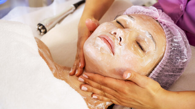 Cosmetologist applies  a moisturizing mask on female face. Woman in a spa salon on cosmetic procedures for facial care.  White woman getting beauty treatment therapy.