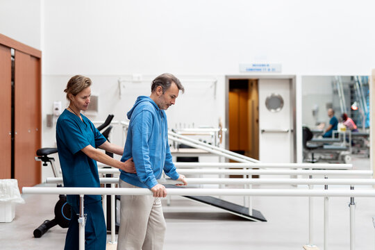 Assistant helping patient to walk between parallel bars