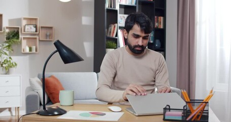 Young Arabian male freelancer ending work at computer, standing up and taking cup of coffee off the table. At home during lockdown. Arab man finishing workday and going away. Cozy room. Homey concept.