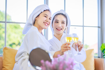 Two Asian young beautiful spa customers friend in white clean bathrobe covered head with towel sitting smiling holding drinking fruit juice beverage glasses talking together after therapy session