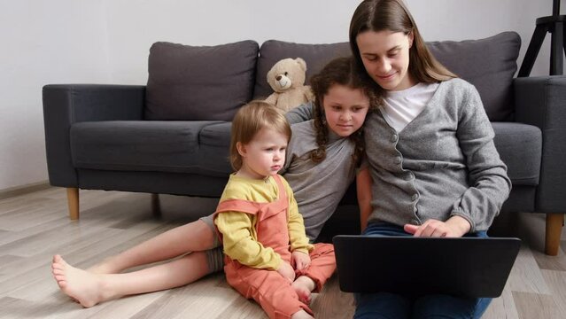 Caring loving young mom with two little daughters kids rest on warm floor at home spend weekend laughing watching family cartoons on laptop, enjoy free time in living room. Modern tech usage concept