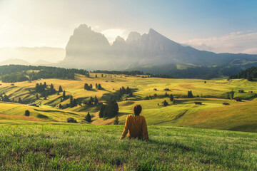 Young man relaxing on alpine meadow in Dolomites Alp mountains Seiser Alm or Alpe di Siusi at sunrise - obrazy, fototapety, plakaty