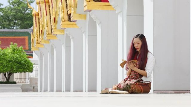 Asian buddhist woman is reading Sanskrit ancient palm leaf manuscript of Tripitaka the Lord Buddha dhamma teaching while sitting in temple on holy full moon day to chant and worship in the monastery