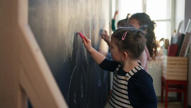 Little girl drawing her friends with chalks on blackboard wall indoors in playroom.