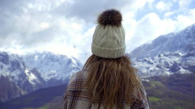 Stylish young girl wears a plaid shirt and a white fleece hat while looking at the views of the picos de europa natural park, some snowy mountains in winter on a sunny day with clouds.