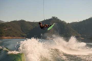 Active sportsman jumping with wakeboard over splashing river wave against the blue sky and green hills