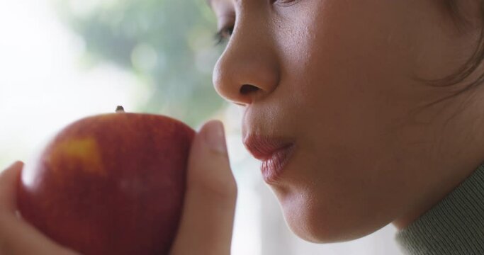 Close-up young cute girl asia people busy work sit at home table hold organic red apple on hand bite full mouth eat fresh fruit snack for weight loss low calories immune vitamin good fiber diet meal. 