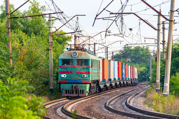 A powerful green locomotive pulls a long train of wagons with containers to the railway station for loading onto a ship. Close-up view.