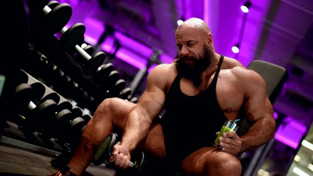 An active athletic male bodybuilder enjoys drinking fresh vitamin juice while sitting on a bench and pumping his biceps