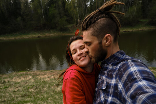 Concept of support and family values. He kisses her on cheek and she smiles. Young European couple man and woman with dreadlocks take selfie in spring park. Funny people hug by river in summer.