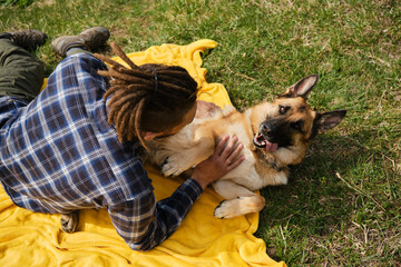 Young Caucasian man with dreadlocks is resting in park with dog. Male owner strokes German Shepherd...