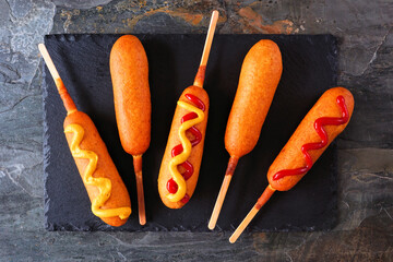 Corn dogs with various toppings on a slate platter. Above view over a dark background.