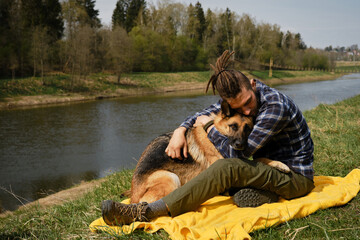 Young Caucasian man with dreadlocks is resting sitting on yellow blanket in park with dog. Owner hugs German Shepherd on green grass on riverbank in spring. Concept of pet as family member.