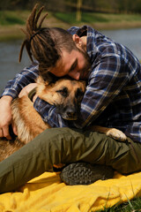 Young Caucasian man with dreadlocks is resting sitting on yellow blanket in park with dog. Owner hugs German Shepherd on green grass on riverbank in spring. Concept of pet as family member.