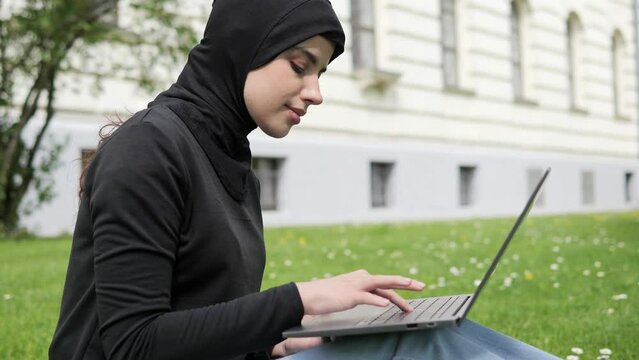 Cute muslim arab islamic student girl in hijab. Woman freelancer user uses laptop sitting on green grass in park near university working online studying remotely chatting browsing shopping outdoor.