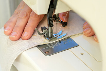 A woman sews on a sewing machine. Women's hands substitute the fabric for sewing under the foot of the sewing machine. - obrazy, fototapety, plakaty