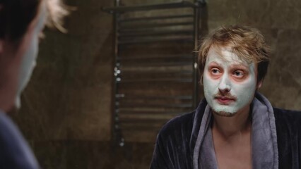A man in a bathrobe applied a cosmetic mask to his face while looking in the mirror in the...
