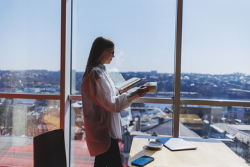 Thoughtful businesswoman with notepad, dressed in formal wear, standing near panoramic window in corporate company and thinking about trade investment, thoughtful businesswoman in glasses