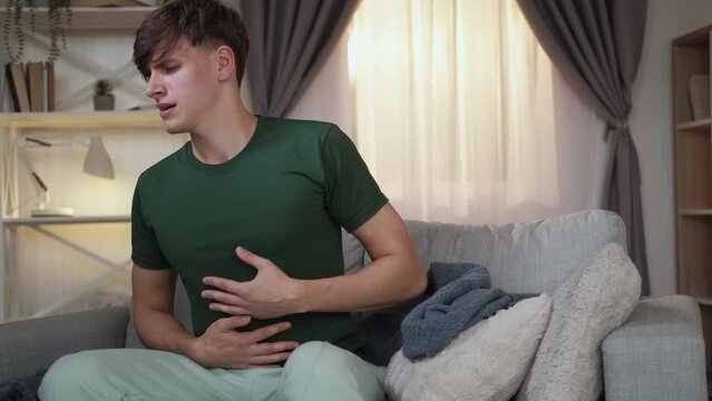 Heavy meal. Suffering man. Stomach pain. Casual guy feeling sick after dinner grasping belly in cramps on sofa light home interior.